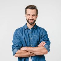 Confident caucasian young man in casual denim clothes with arms crossed looking at camera with toothy smile isolated in white background